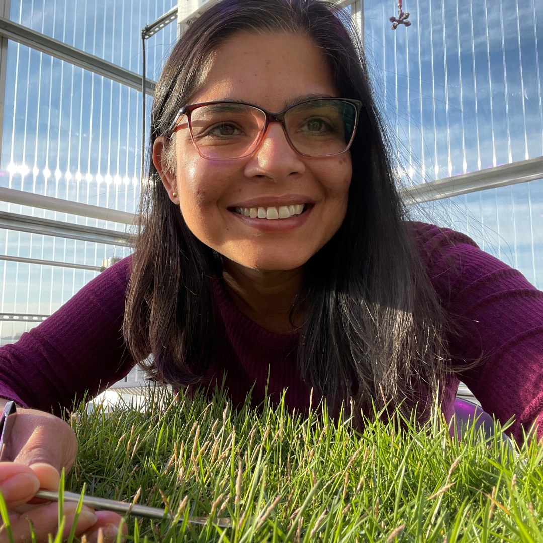 Portrait of a woman smiling over a tray of turfgrass in a greenhouse wearing glasses 